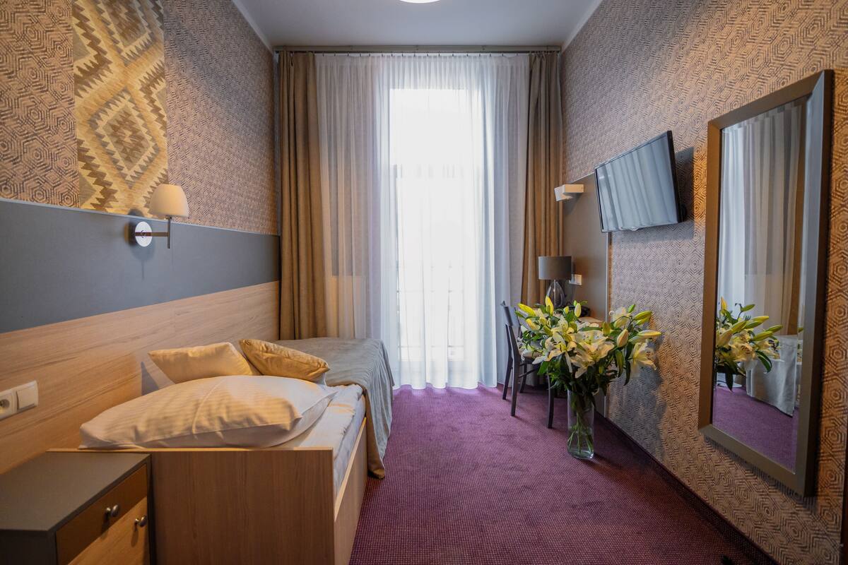 A hotel room with a bed, bedside table, lamp, mirror, and a desk with a chair.