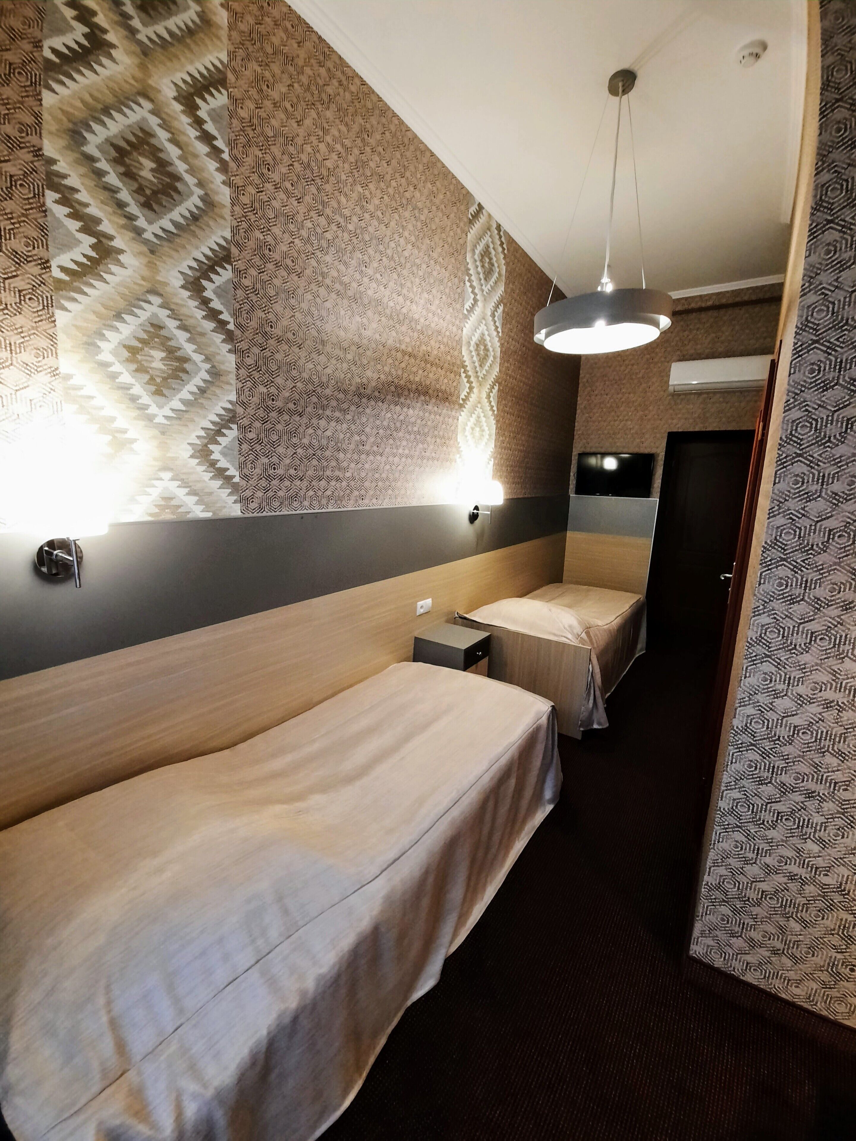 A hotel room with two single beds, a bedside table, a wall-mounted TV, and a patterned wall design.