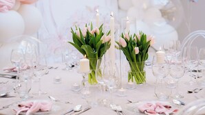 Una mesa redonda dispuesta para un evento formal, con copas transparentes, velas y servilletas rosas.