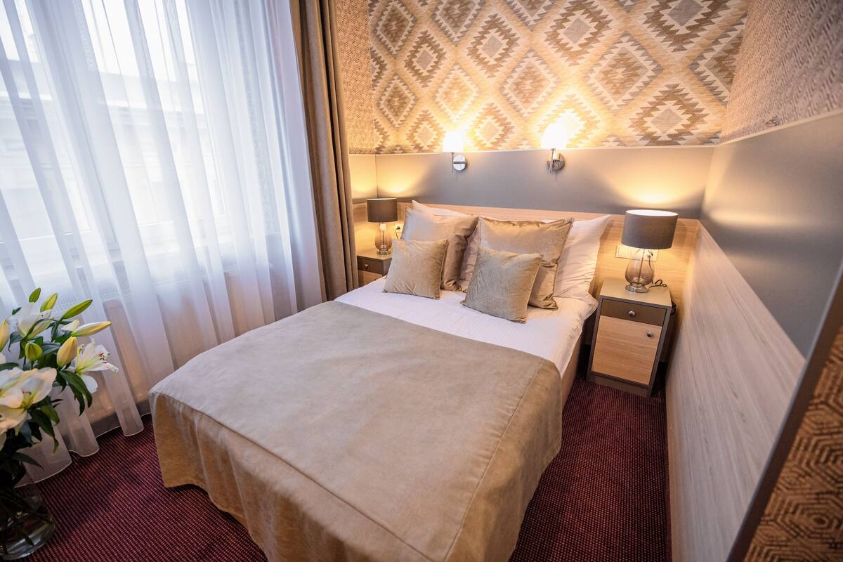 A hotel room with a large bed, two bedside tables with lamps, a patterned wall, and sheer curtains.