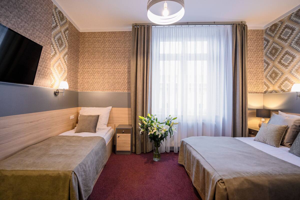 A hotel room with two beds, a TV, a vase of flowers, and a large window with curtains.