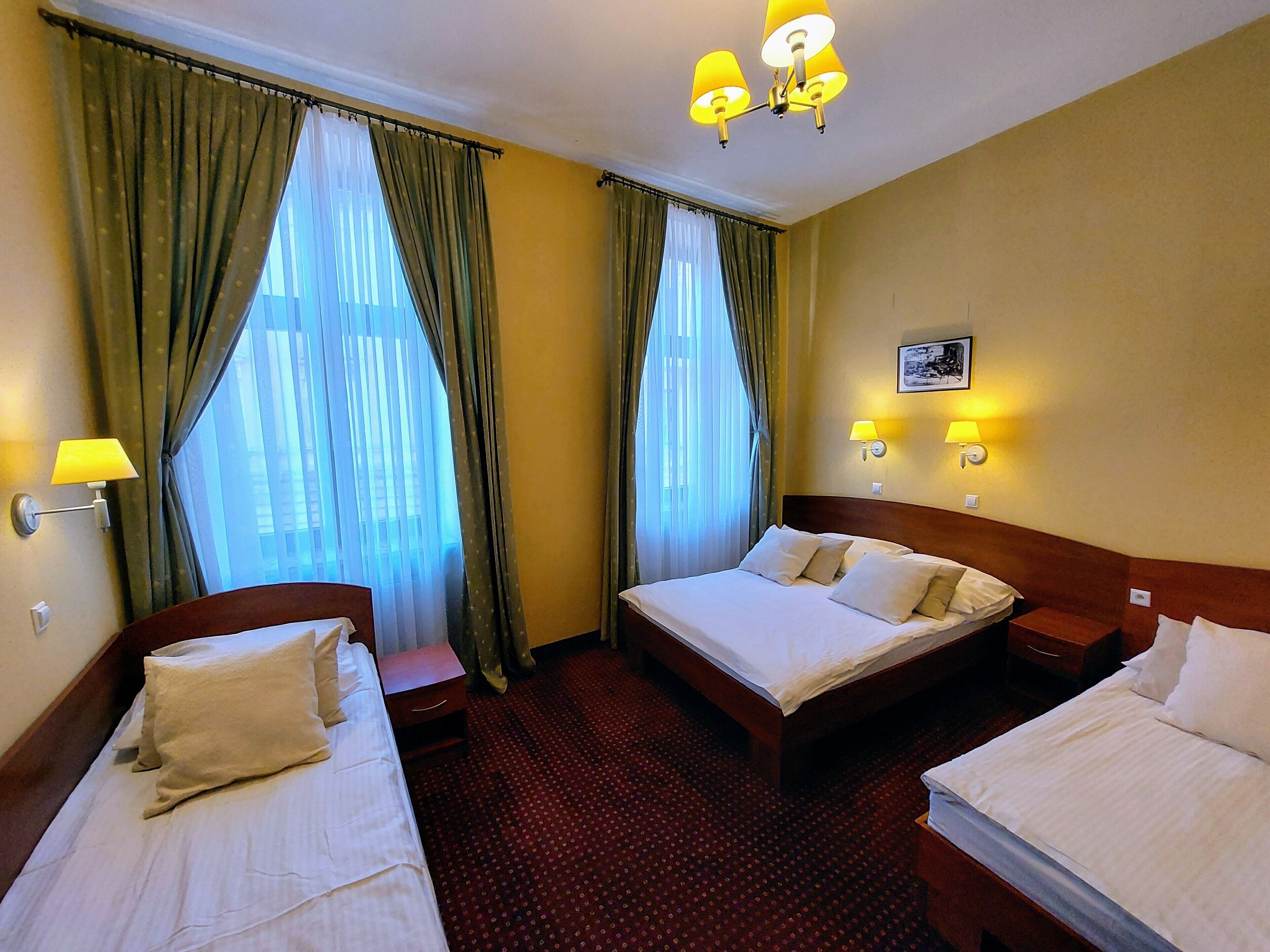 A hotel room with two single beds, a large window with curtains, a chandelier, and wall-mounted lamps.