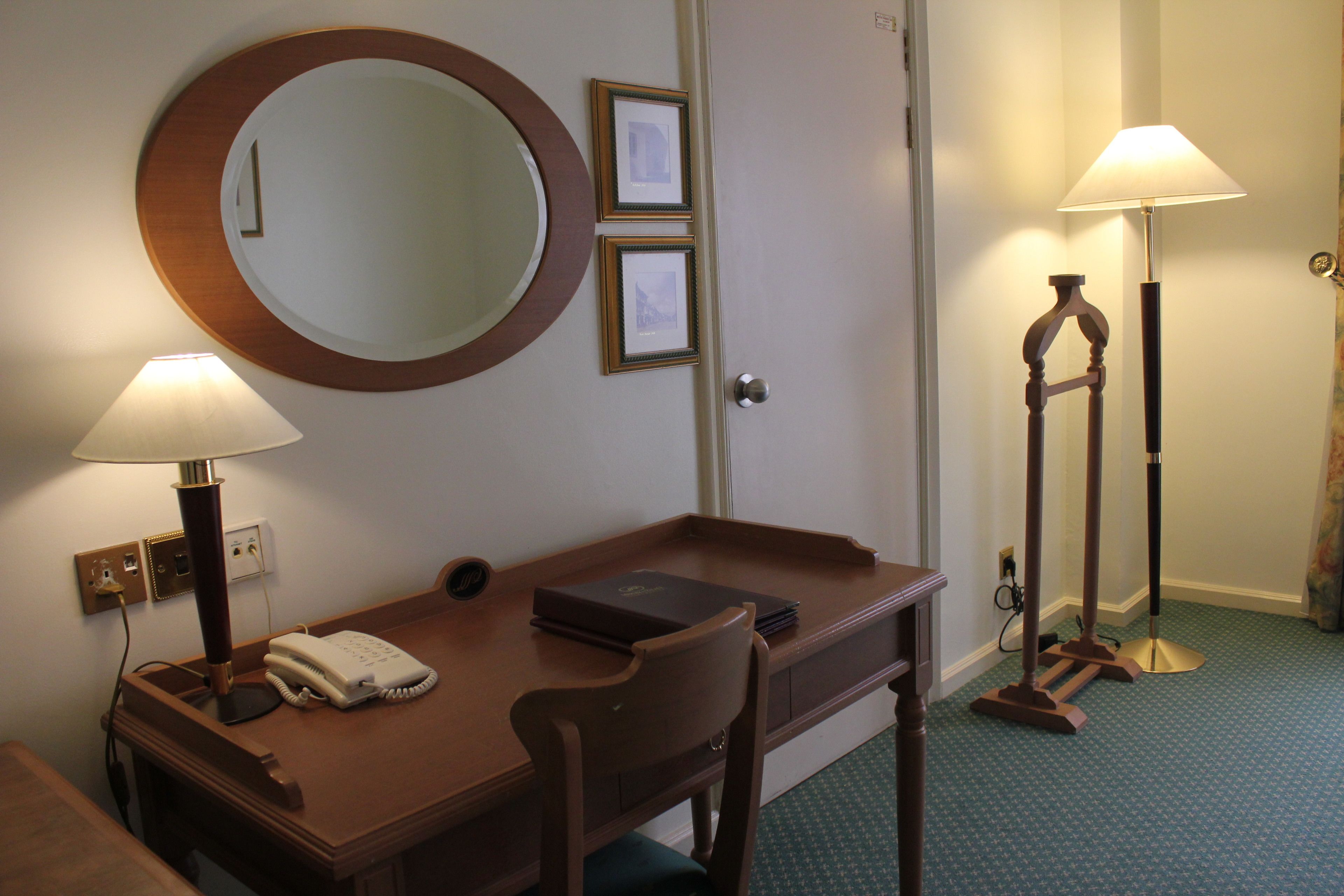 A hotel room with a wooden desk, a telephone, a lamp, a mirror, and a floor lamp.