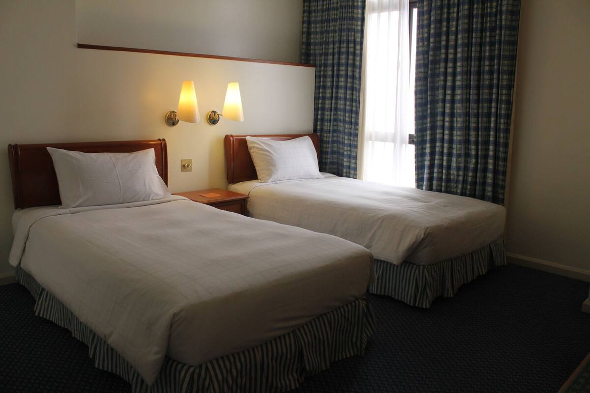 A hotel room with two single beds, a nightstand, a wall-mounted lamp, and a window with curtains.