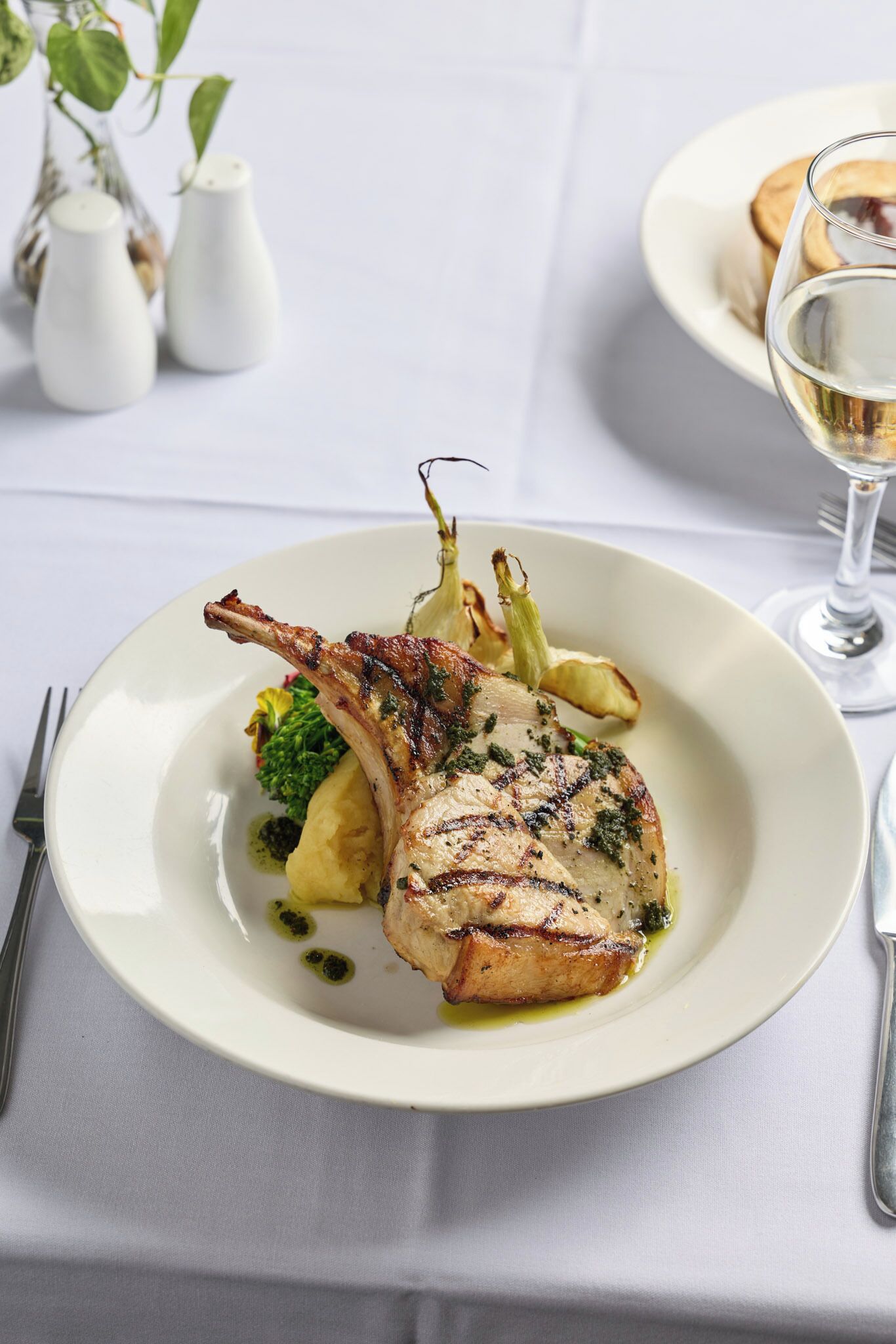A plate of food with a lamb chop, vegetables, and a glass of white wine on a table.
