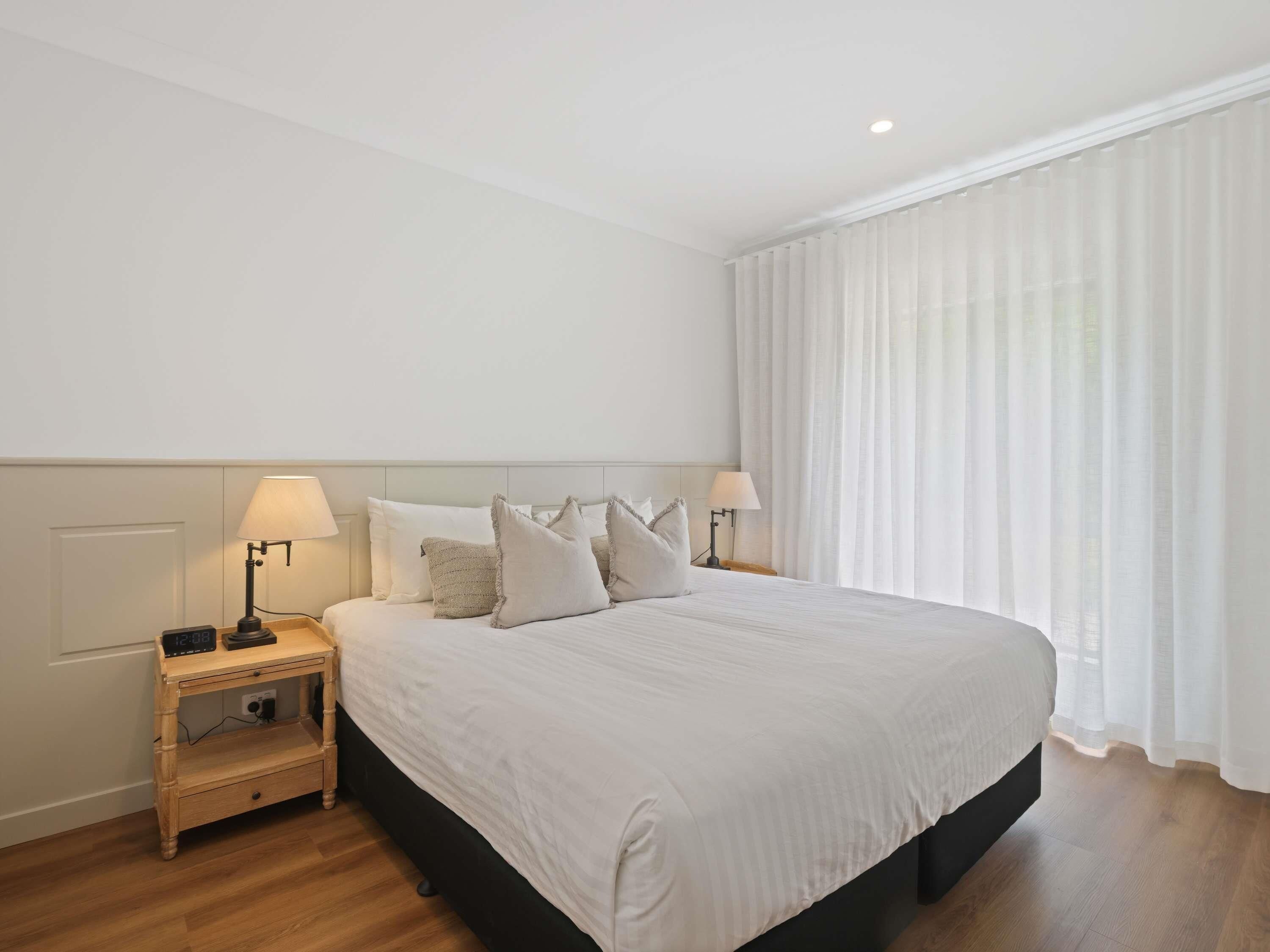 Una cama grande con ropa de cama blanca, dos mesitas de noche con lámparas, un tocador de madera y cortinas blancas traslúcidas.