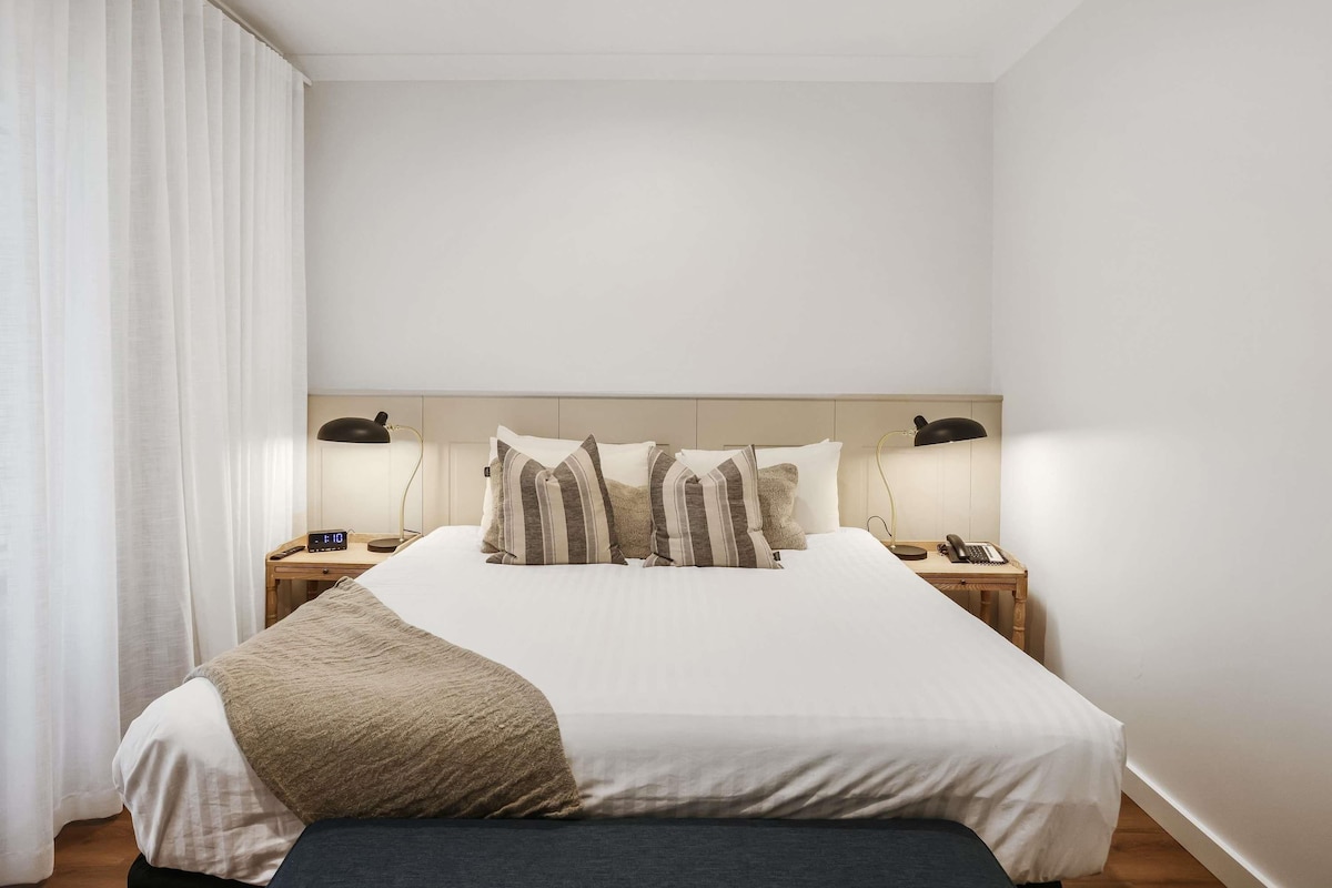 A hotel room with a large bed, two bedside tables with lamps, a clock, and a window with curtains.