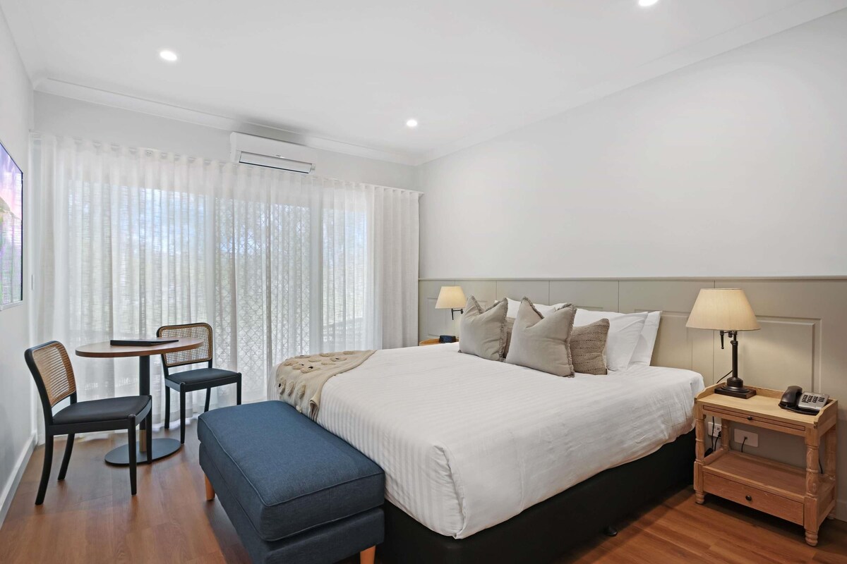A hotel room with a bed, bedside tables, a desk with a chair, and a window with curtains.