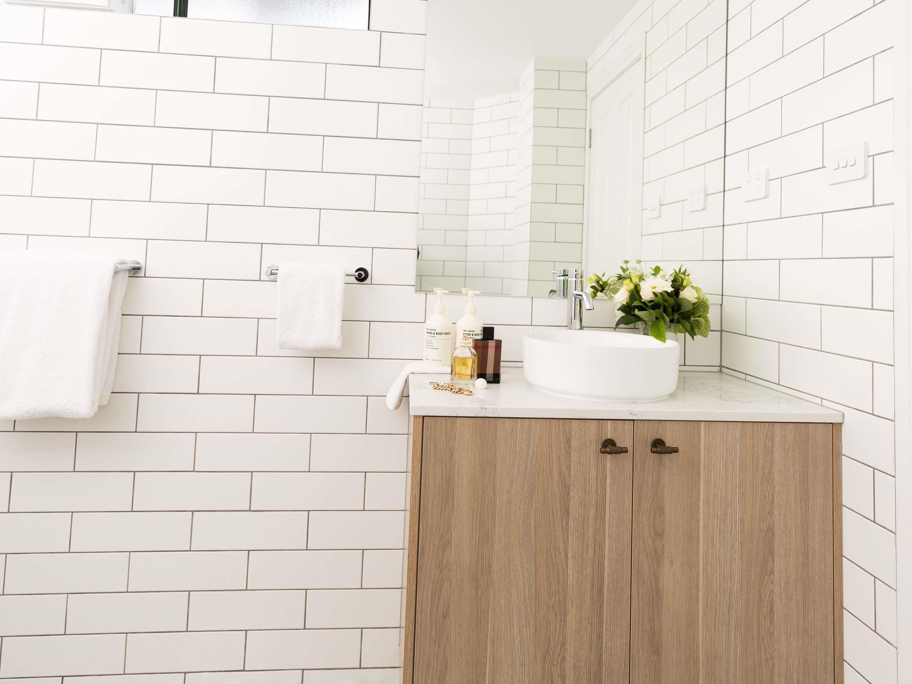 A bathroom with white subway tiles, a white sink, a wooden cabinet, and a mirror.