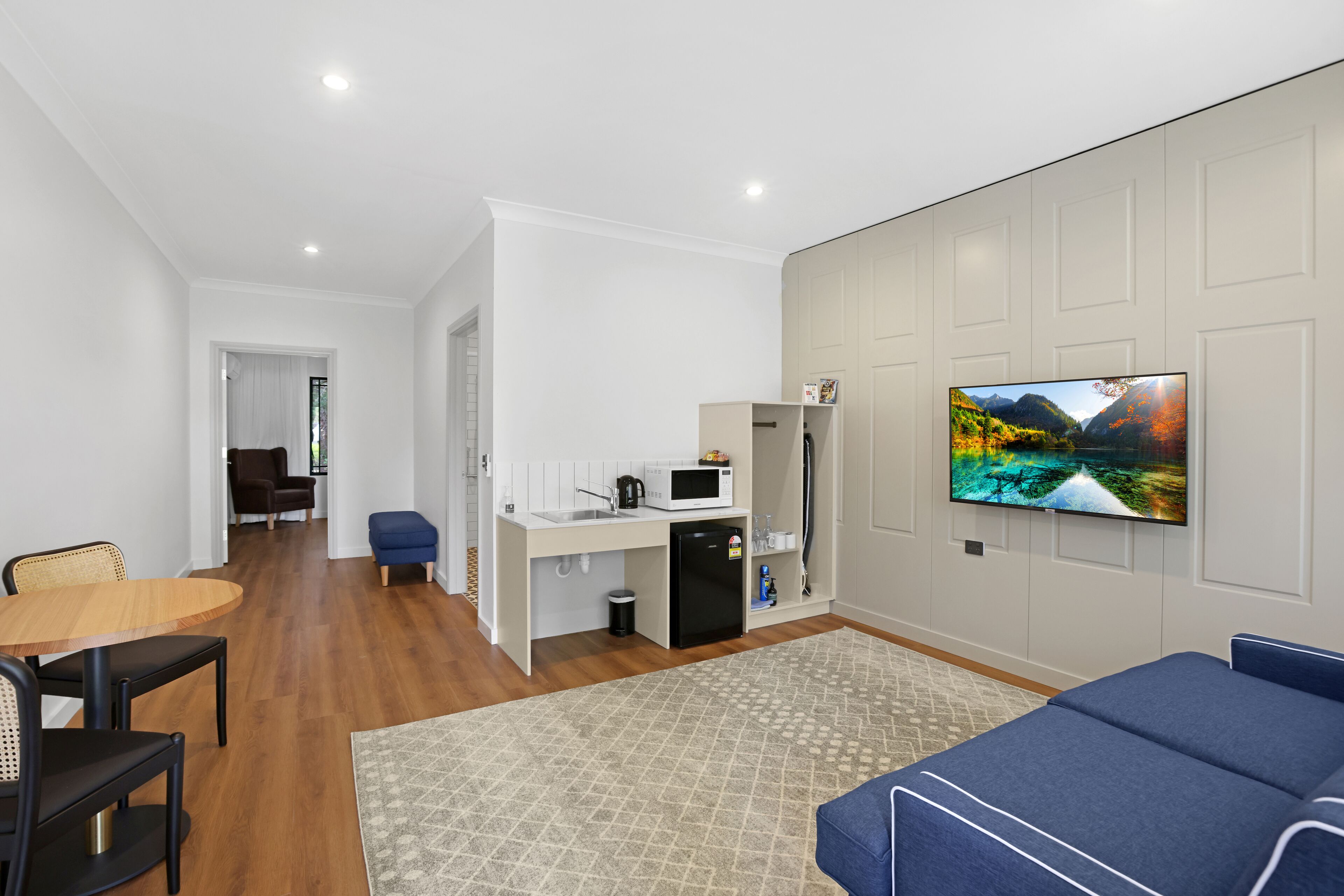 Een moderne woonkamer met een flatscreen televisie, een eethoek met een houten tafel en stoelen, een kitchenette met een magnetron en koelkast, en een kamerbreed tapijt met een patroon.