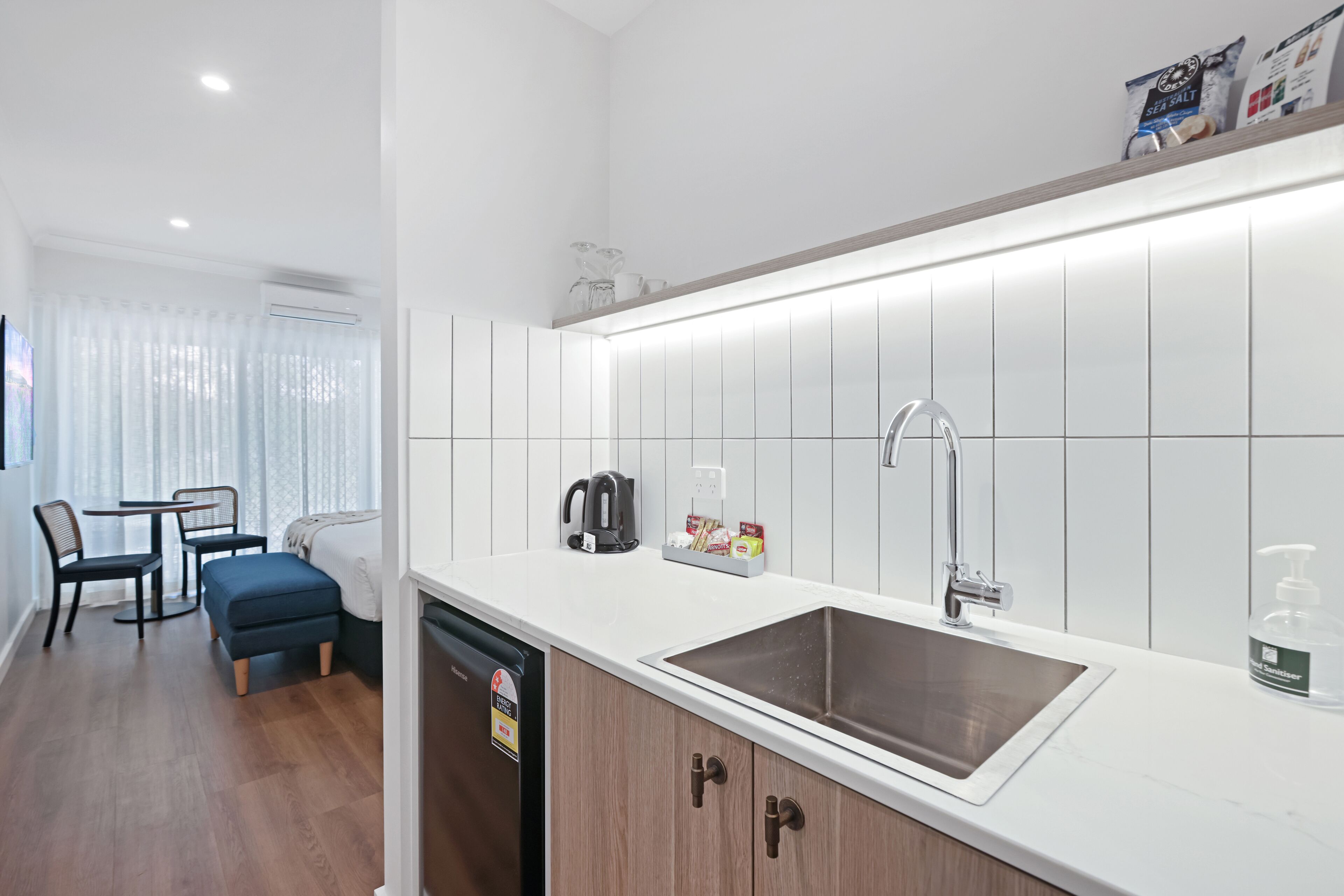 A modern kitchen with a white countertop, stainless steel sink, and wooden cabinets. There is a dining area with a table and chairs, and a window with curtains.