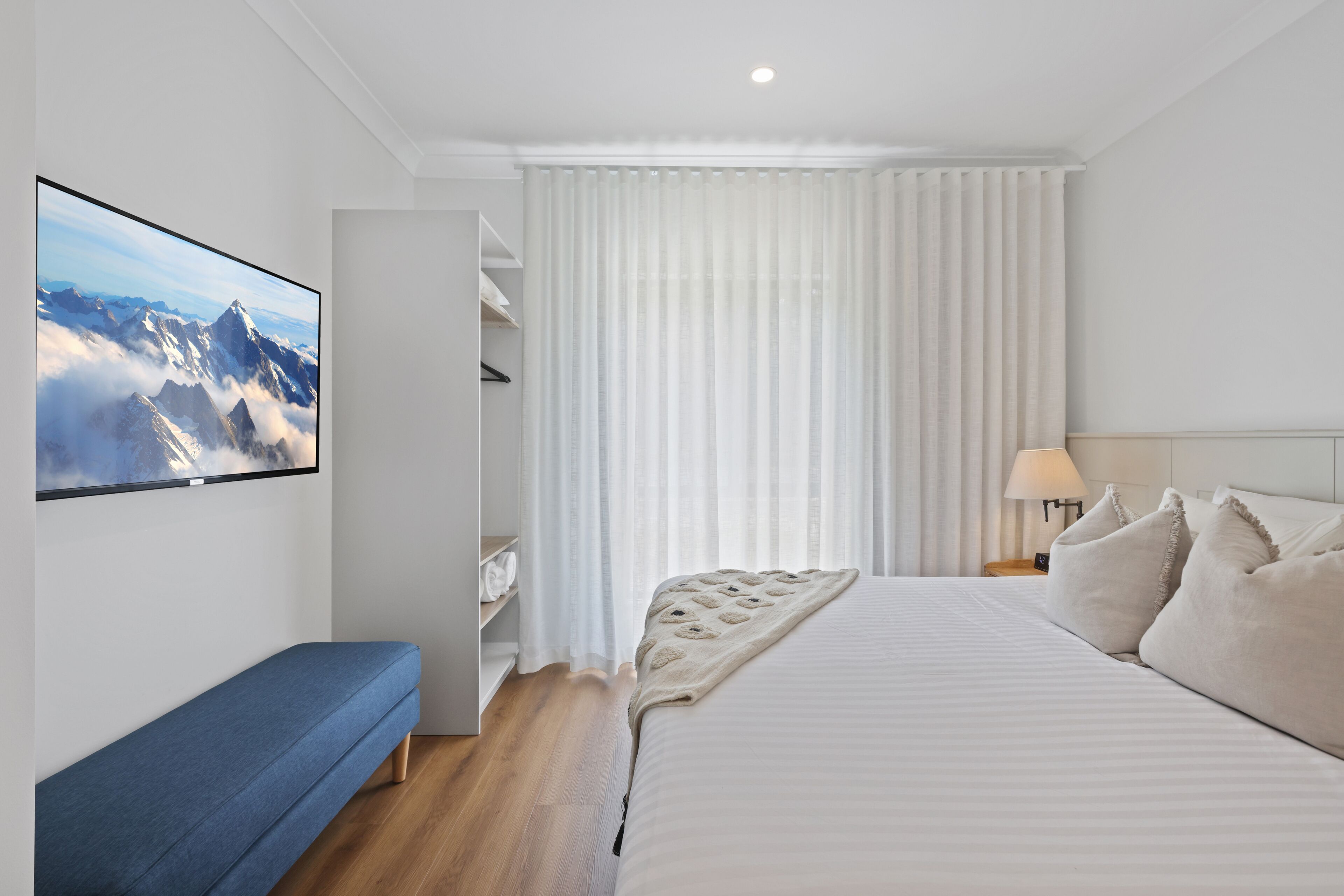A modern hotel room with a large bed, a bench, a wardrobe, and a view of mountains through sheer curtains.