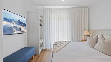 Una habitación de hotel moderna con una cama grande, un banco, un armario y vistas a las montañas a través de cortinas translúcidas.
