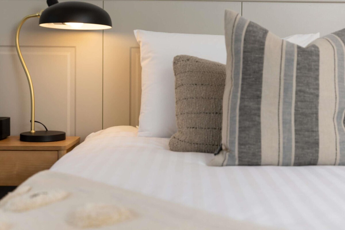 A neatly made bed with white bedding, a striped pillow, and a round, light-colored pillow. A black desk lamp with a gold base is on the nightstand beside the bed.