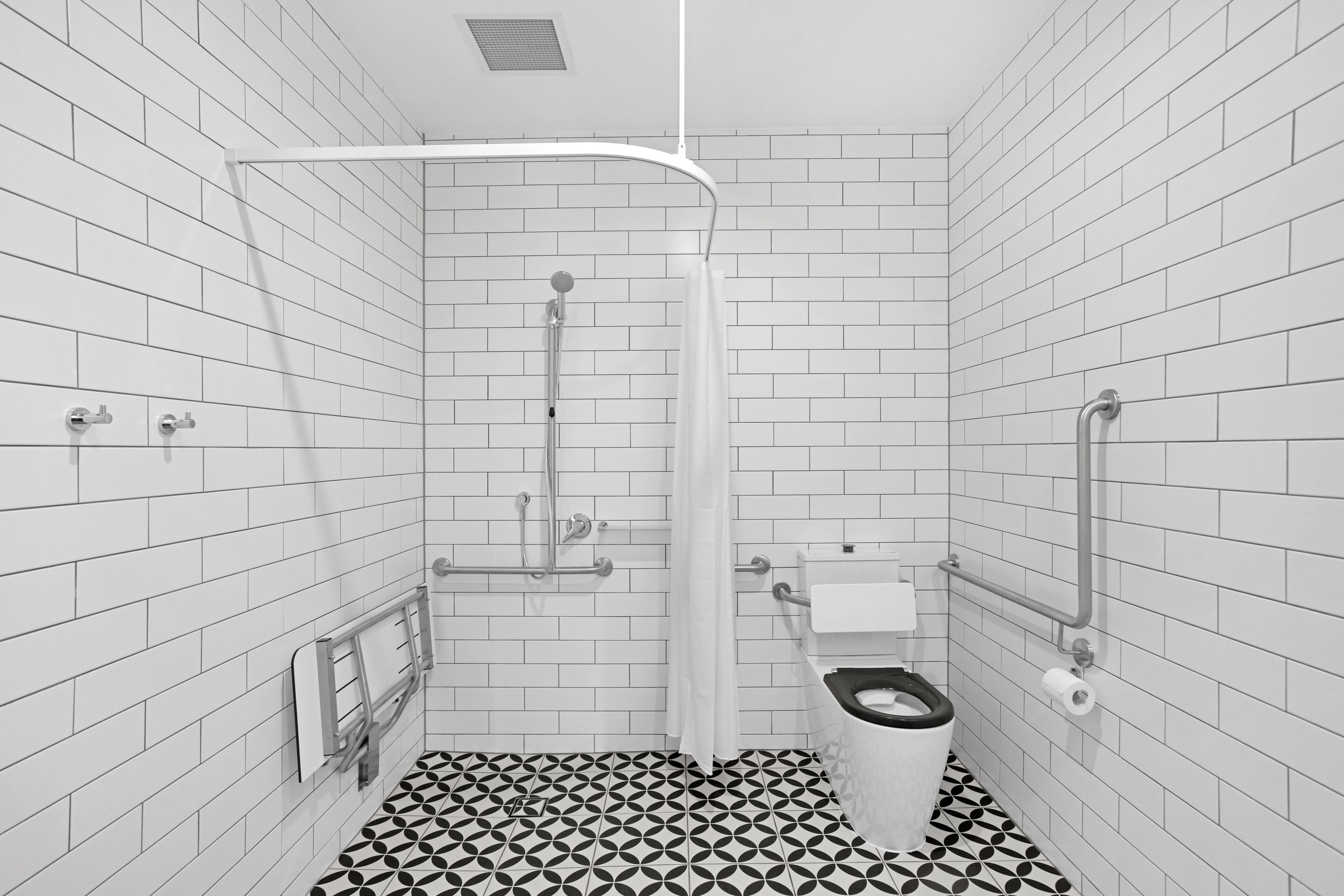 A small, modern bathroom with white tiled walls and floor, a black and white patterned tile floor, a shower, a toilet, and a sink.