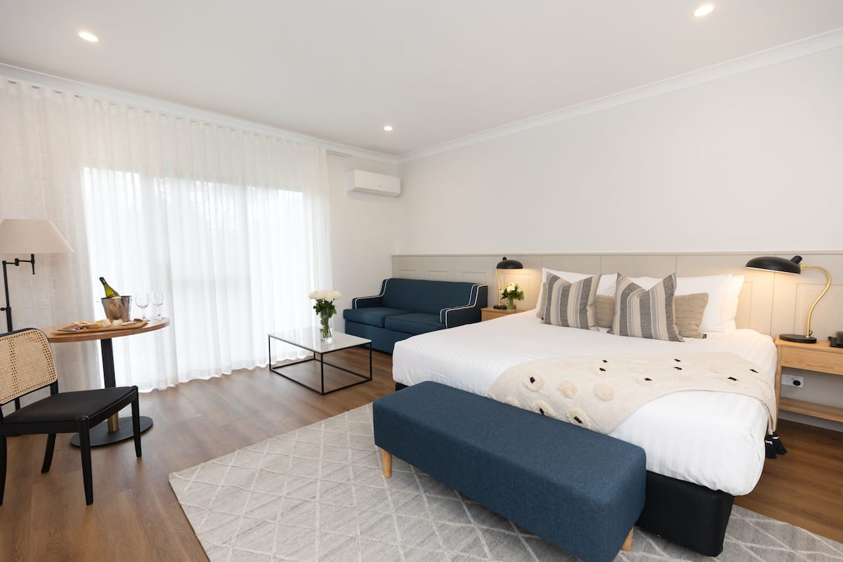 A modern hotel room with a large bed, a small desk, a sofa, and a dining table.