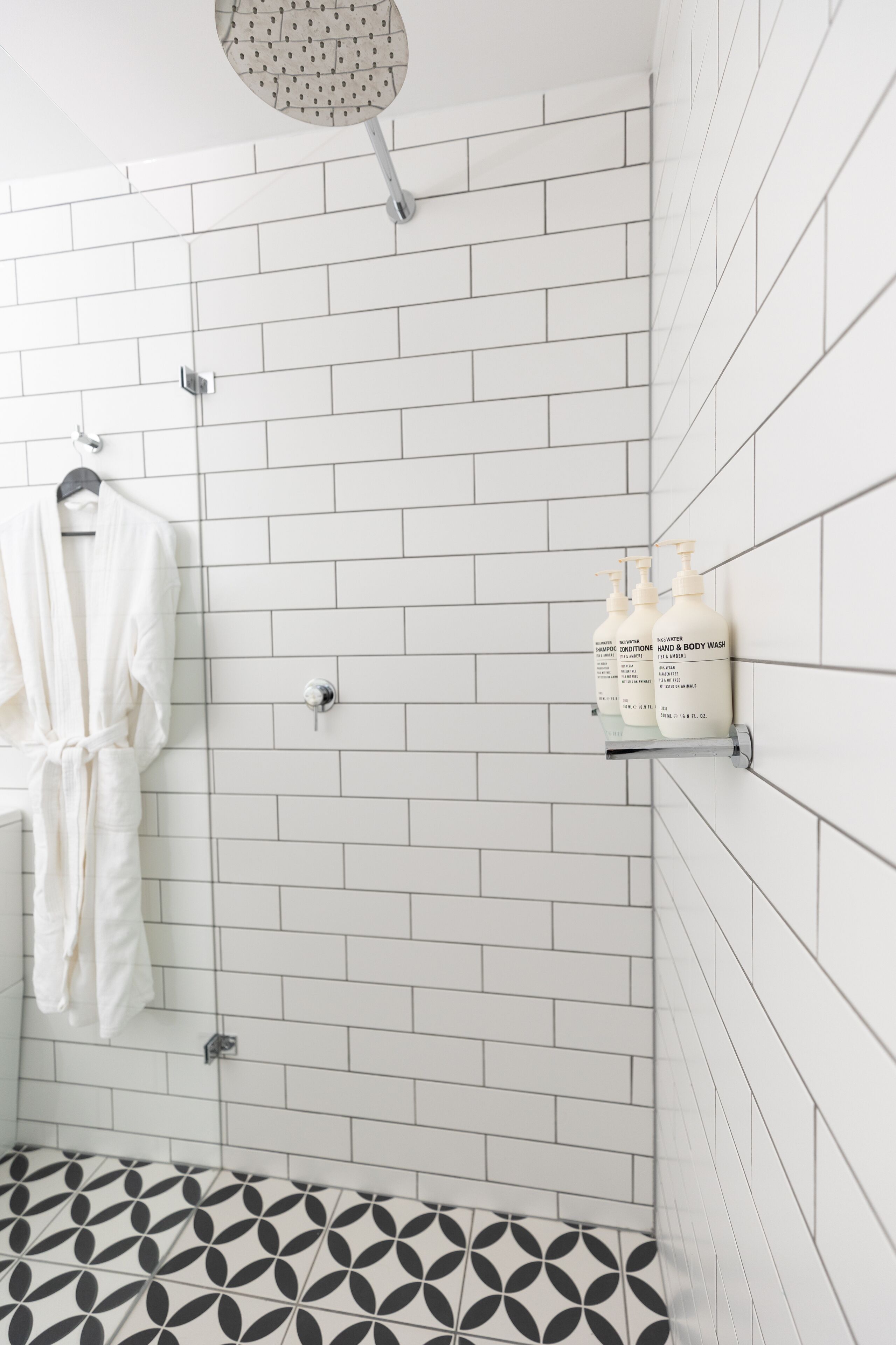A bathroom with white tiled walls, a shower with a handheld showerhead, a hanging bathrobe, and two bottles of hand and body wash on a shelf.