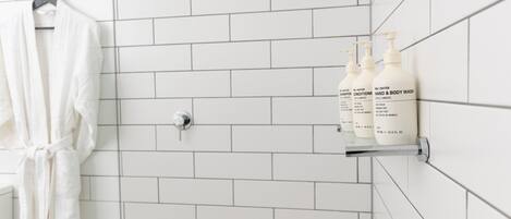 A bathroom with white tiled walls, a shower with a handheld showerhead, a hanging bathrobe, and two bottles of hand and body wash on a shelf.