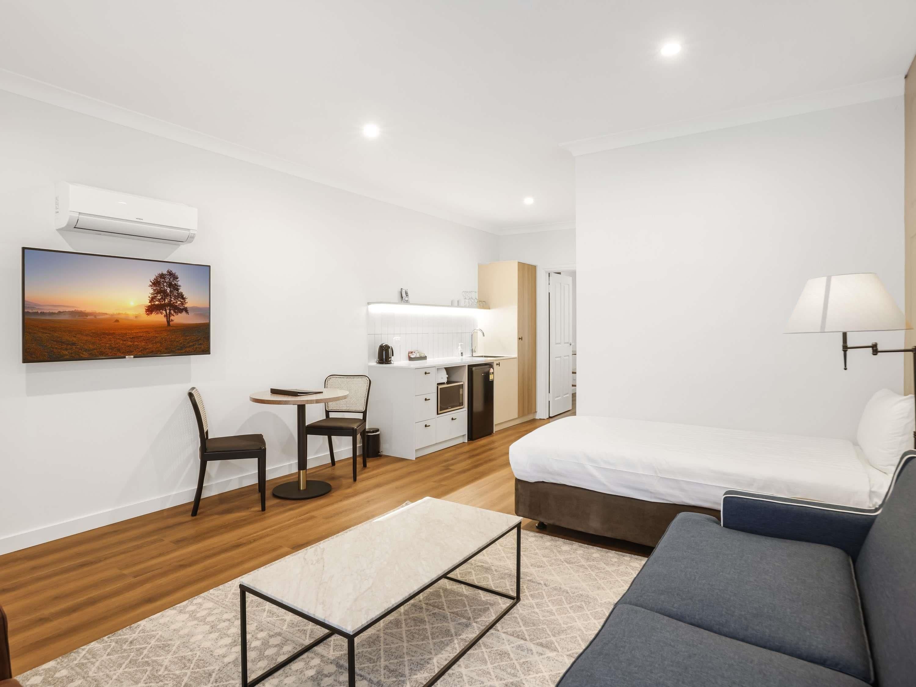 Una habitación moderna y luminosa con una cama, un sofá, una mesita, una silla, un escritorio con silla, un televisor montado en la pared y una ventana.