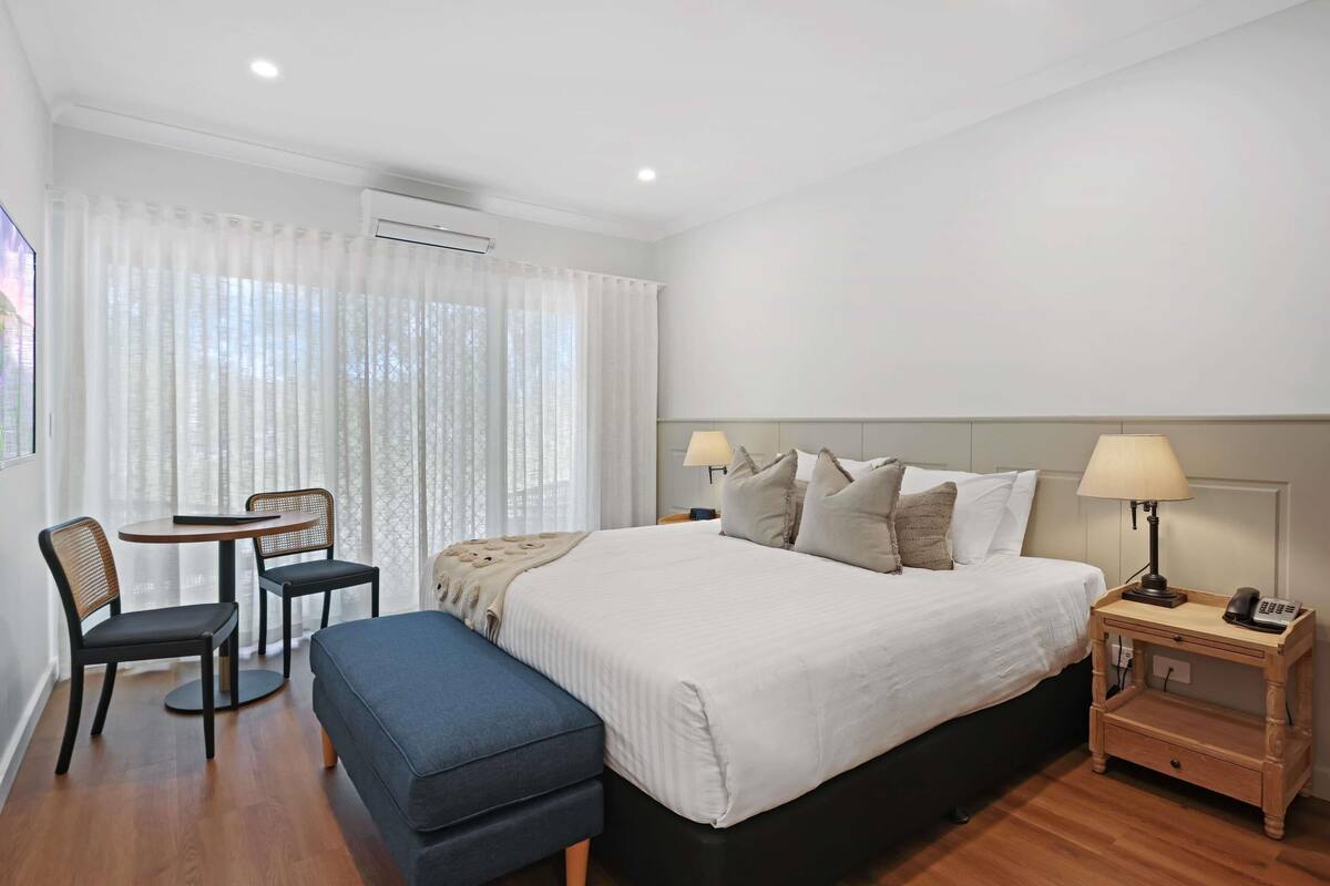 A hotel room with a bed, bedside tables, a desk with a chair, and a window with curtains.
