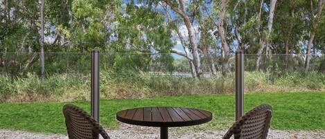 A round wooden table with two wicker chairs, set on a gravel surface, with a view of a grassy area and trees in the background.