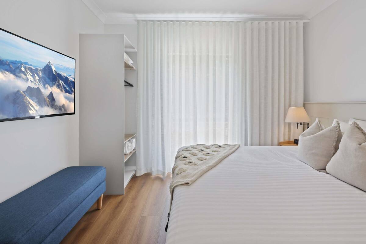 A bedroom with a bed, a bench, a wardrobe, and a TV displaying a mountain view.