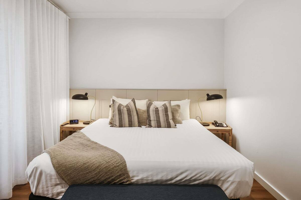 A hotel room with a large bed, two bedside tables with lamps, a clock, and a window with curtains.