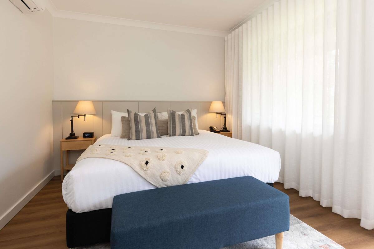 A neatly arranged hotel room with a large bed, two bedside tables with lamps, a bedside clock, and a window with sheer curtains.
