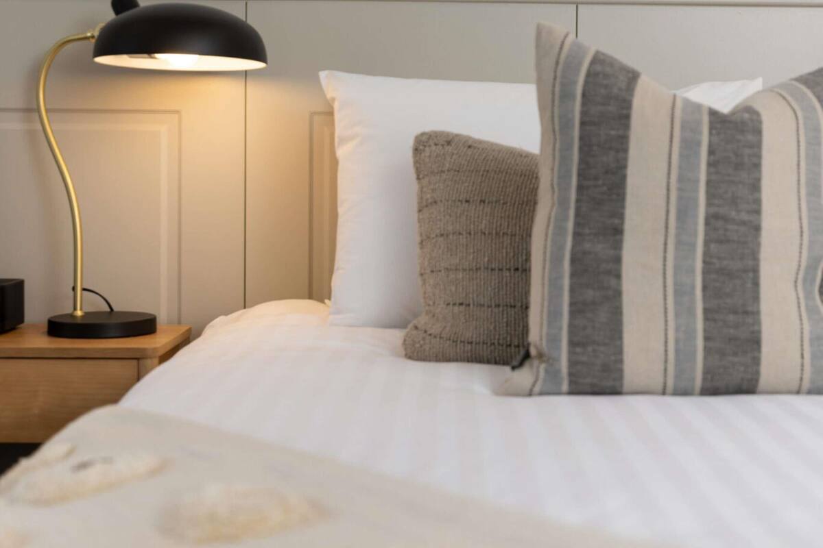 A neatly made bed with white bedding, a striped pillow, and a round, light-colored pillow. A black desk lamp with a gold base is on the nightstand beside the bed.