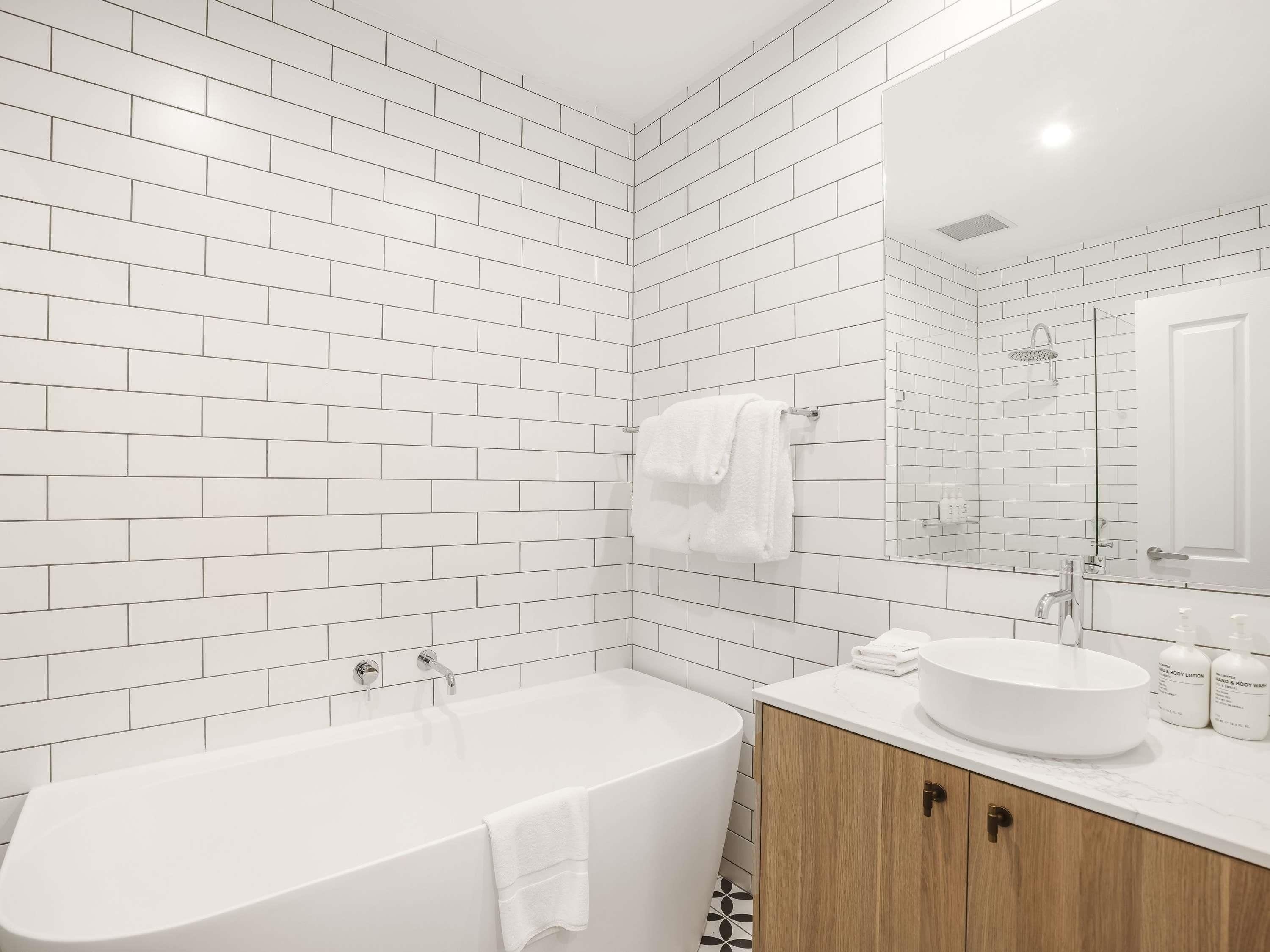 A modern bathroom with white subway tiles, a freestanding bathtub, a wooden vanity with a round sink, and a large mirror.