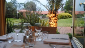 A dining table set with wine glasses, cutlery, and a napkin, with a view of a garden and a building in the background.