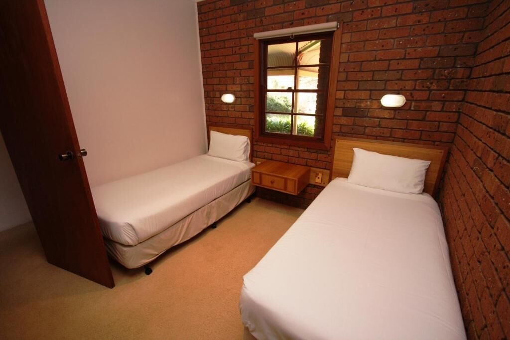 A small room with two single beds, a wooden nightstand, and a window with a view of greenery.