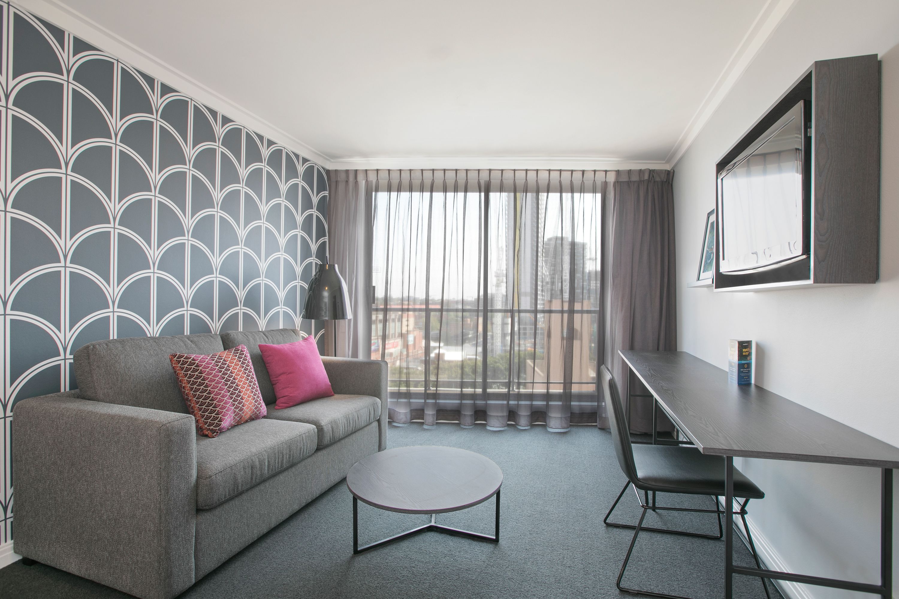 Ein modernes Wohnzimmer mit grauem Sofa, rundem Couchtisch und Esstisch mit Stühlen. Ein großes Fenster mit transparenten Vorhängen bietet Blick auf die Stadt.