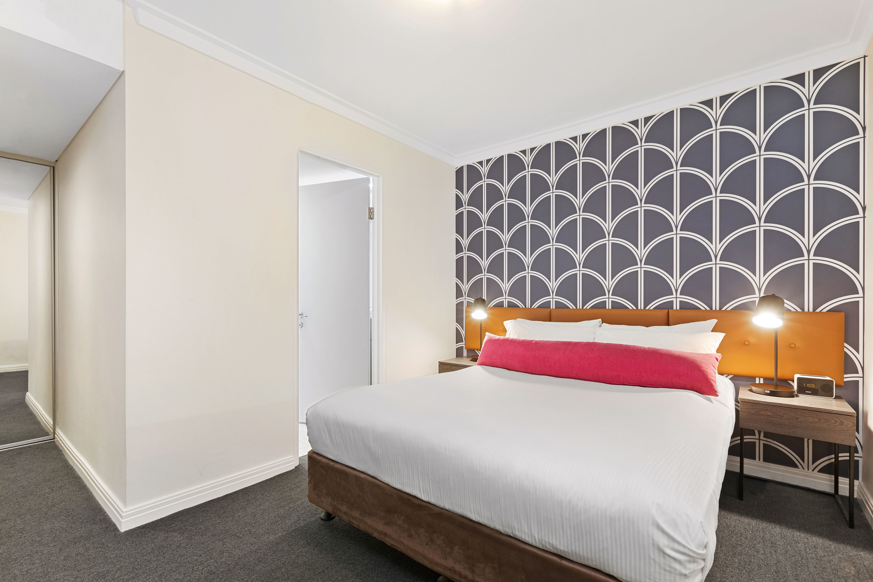 A hotel room with a large bed, a nightstand with a lamp, and a patterned wallpaper.
