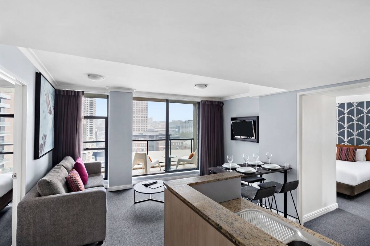 A modern hotel room with a sofa, dining table, chairs, a TV, and a balcony with a city view.