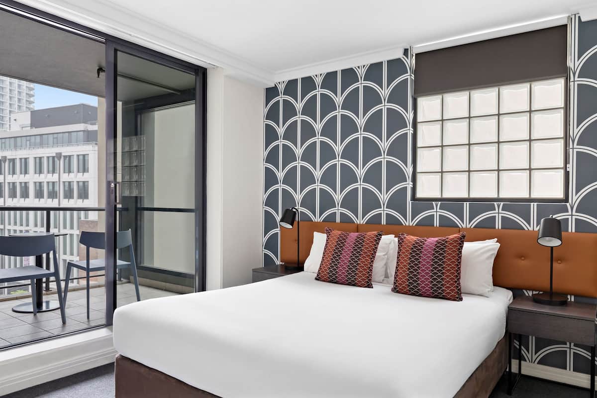 A hotel room with a large bed, a balcony with chairs, a patterned wall, and a window with blinds.