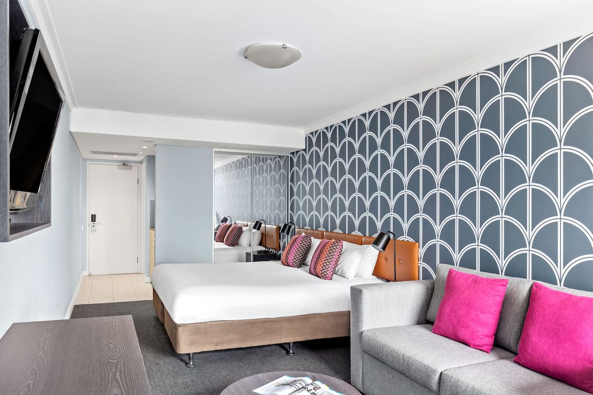 A modern hotel room with a large bed, a grey sofa, and a TV mounted on the wall.
