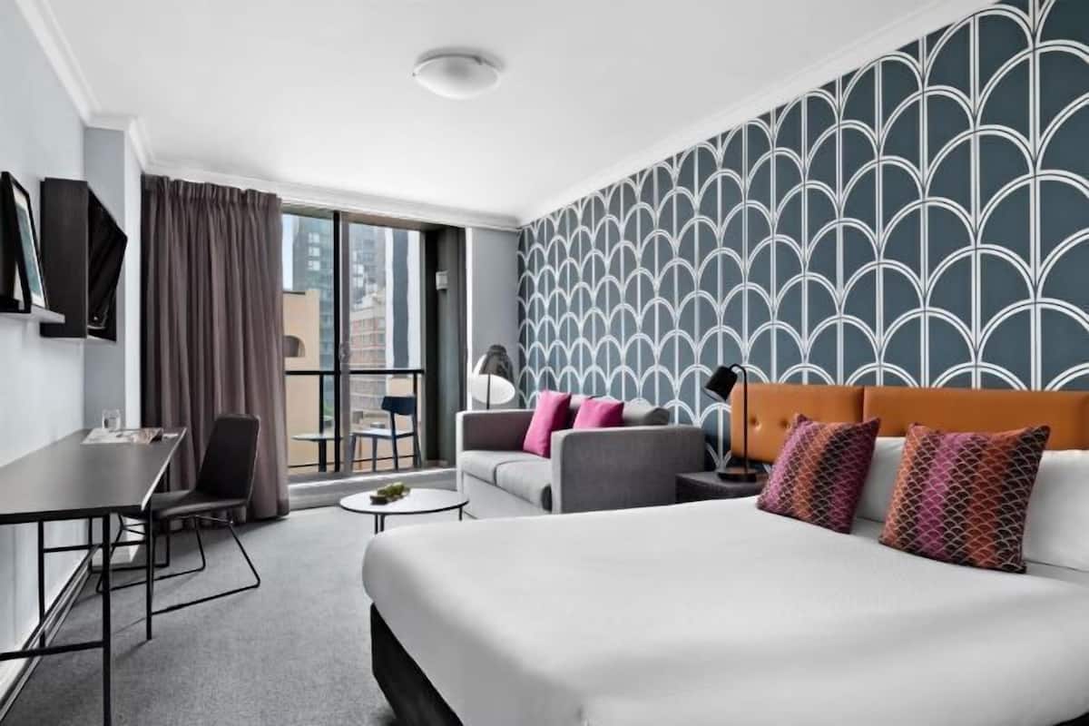 A hotel room with a large bed, a sofa, a desk, and a view of the city through the window.