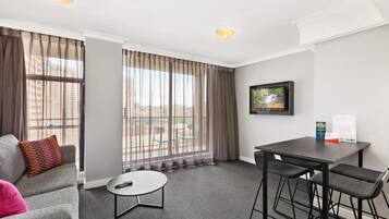 A hotel room with a grey sofa, a small round table, two bar stools, a TV on the wall, and a large window with sheer curtains.