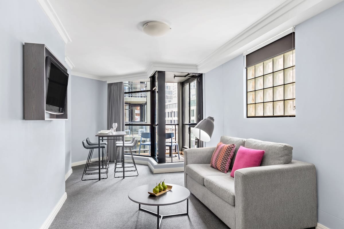 A modern living room with a grey sofa, a coffee table with pears, a TV mounted on the wall, and a large window with a view of the city.