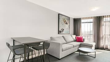 A modern living room with a grey sofa, a small round table, and a dining table with chairs. There is a painting on the wall and a window with curtains.