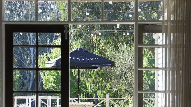 Une terrasse en bois avec un parasol, une clôture blanche et une vue sur la verdure, visible à travers de grandes fenêtres.