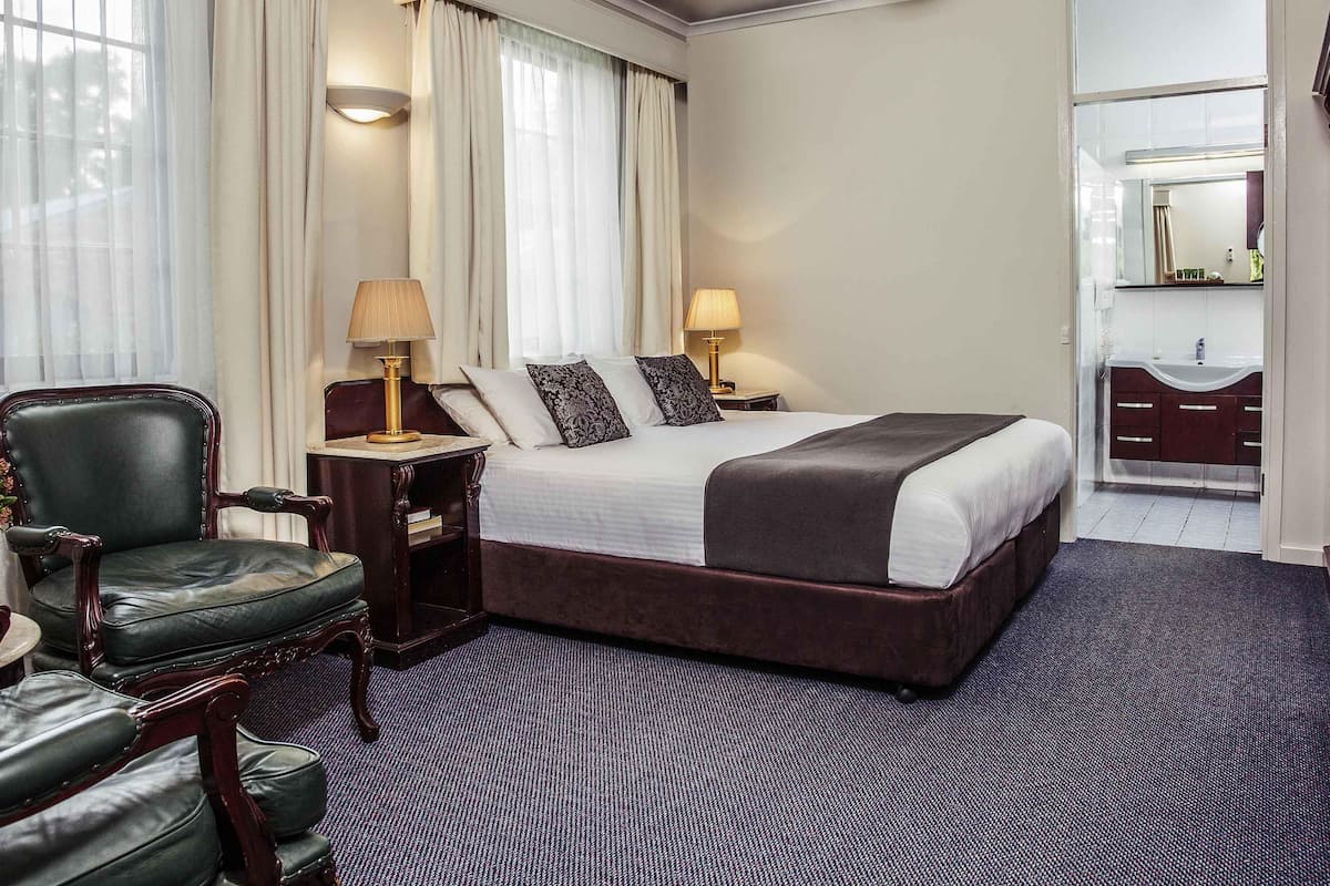 Quarto de hotel com uma cama grande, uma escrivaninha, uma cadeira, toucador com espelho e uma janela com cortinas.