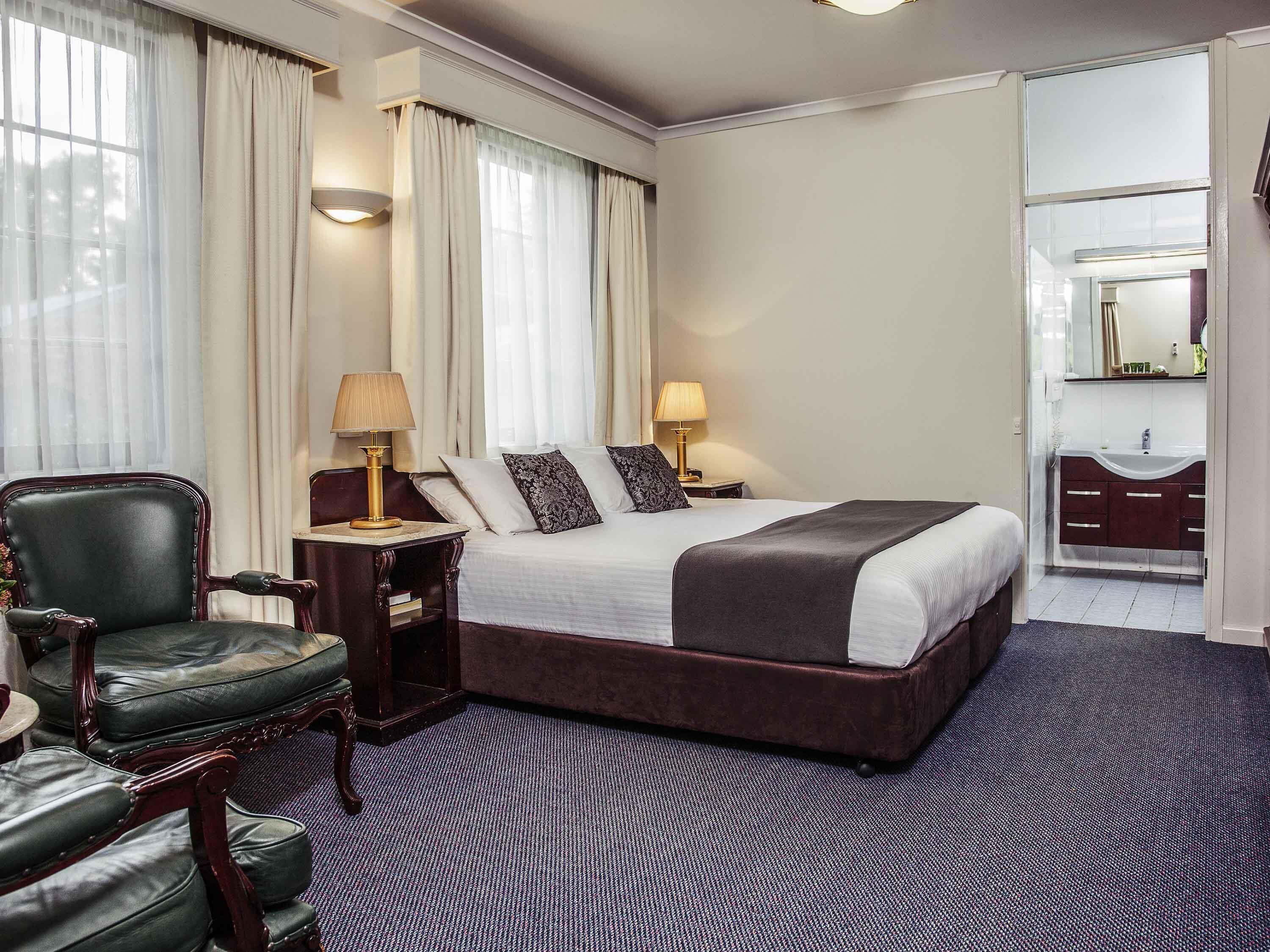 A hotel room with a large bed, a desk, a chair, a vanity with a mirror, and a window with curtains.