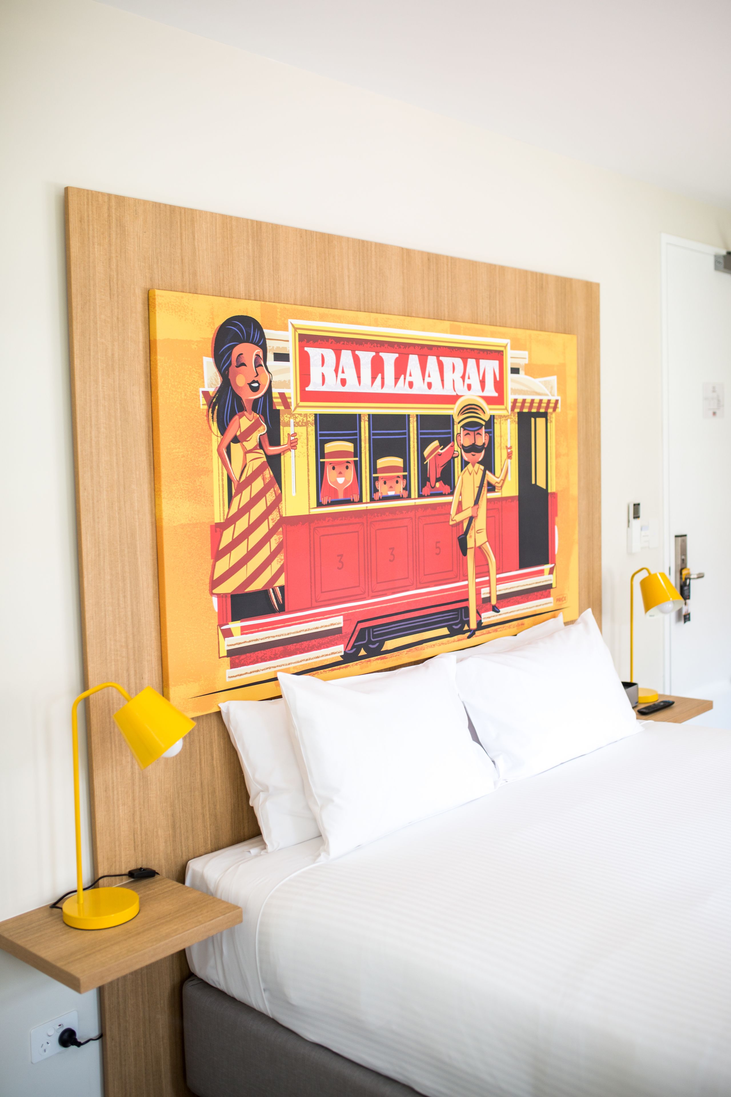 Un dormitorio con cama, almohadas blancas, lámpara de noche amarilla y una obra de arte en la pared que representa un tren antiguo y personajes.
