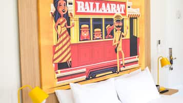 Un dormitorio con cama, almohadas blancas, lámpara de noche amarilla y una obra de arte en la pared que representa un tren antiguo y personajes.