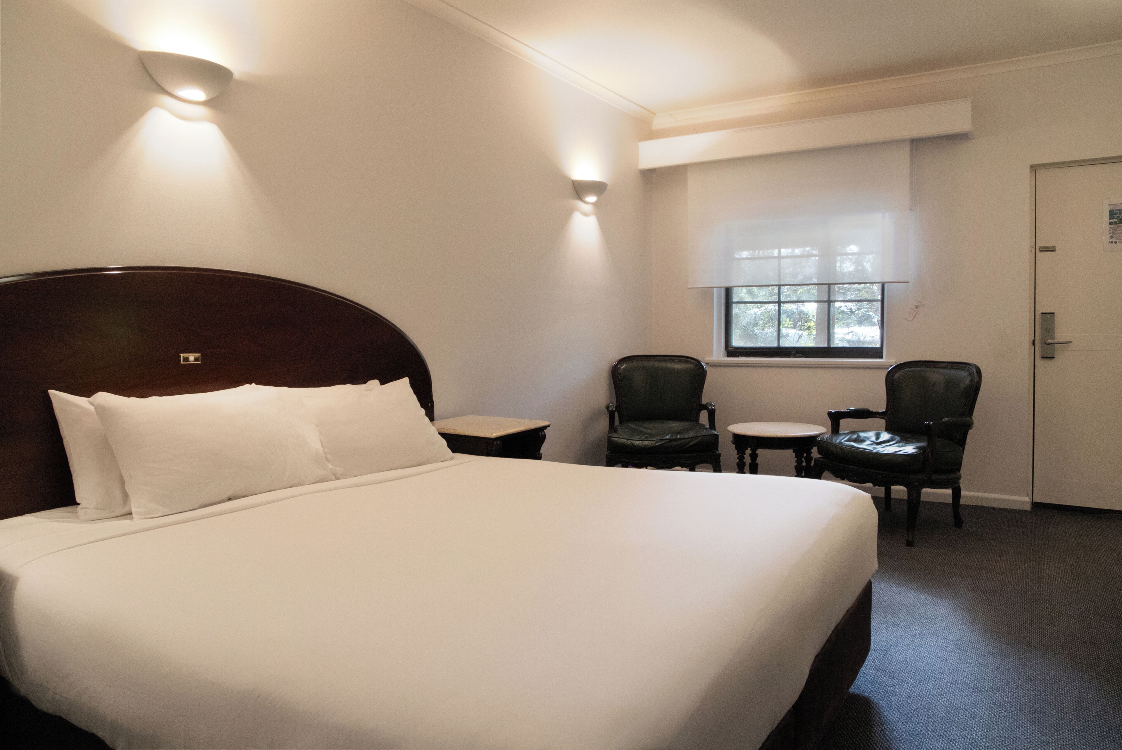 Une chambre d’hôtel avec un grand lit, deux chaises, une petite table et une fenêtre avec des stores.