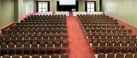 Un salón de conferencias con filas de sillas, un escenario y una pantalla grande.