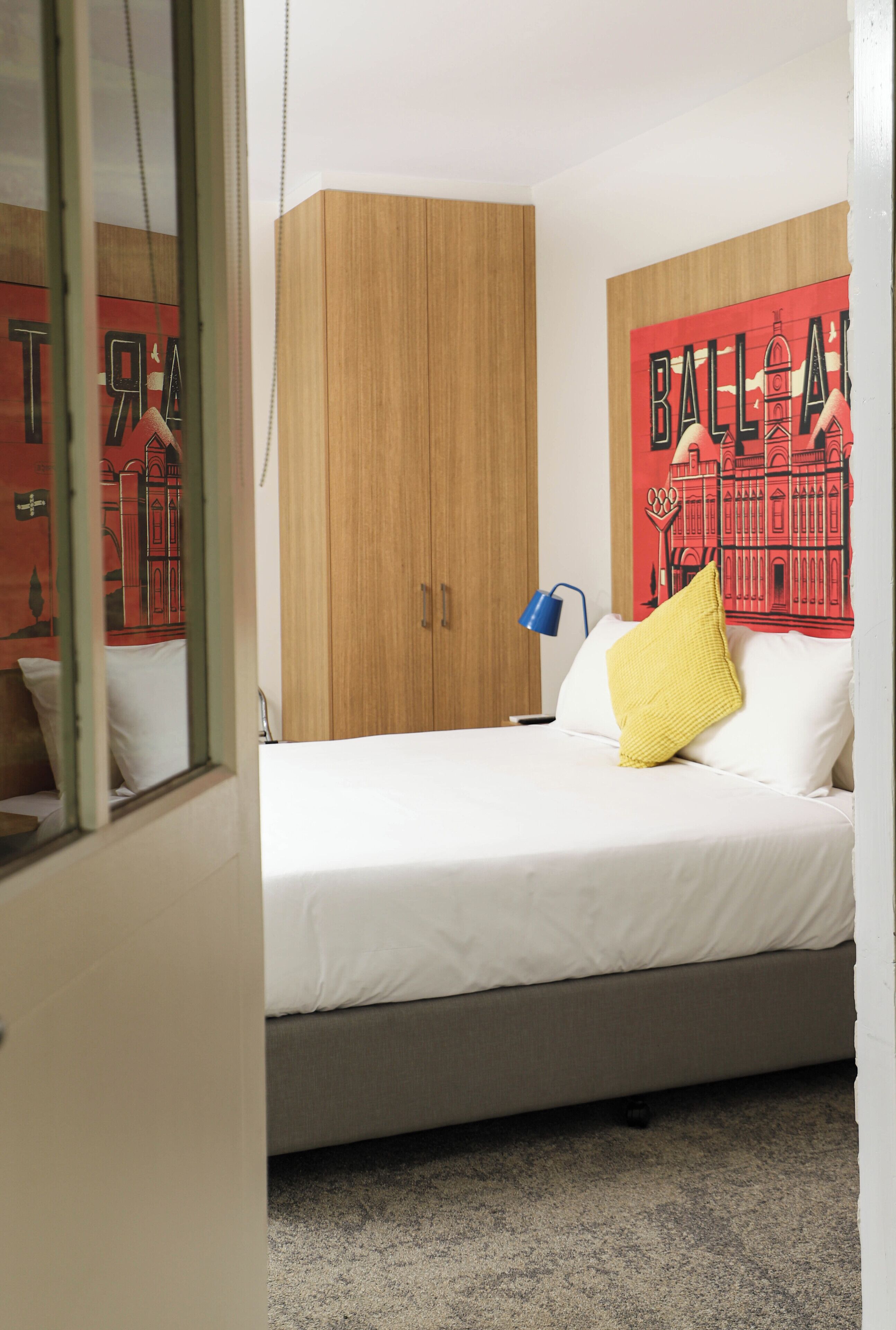 A bedroom with a bed, a wooden wardrobe, a wall with a red and black mural, and a blue bedside lamp.