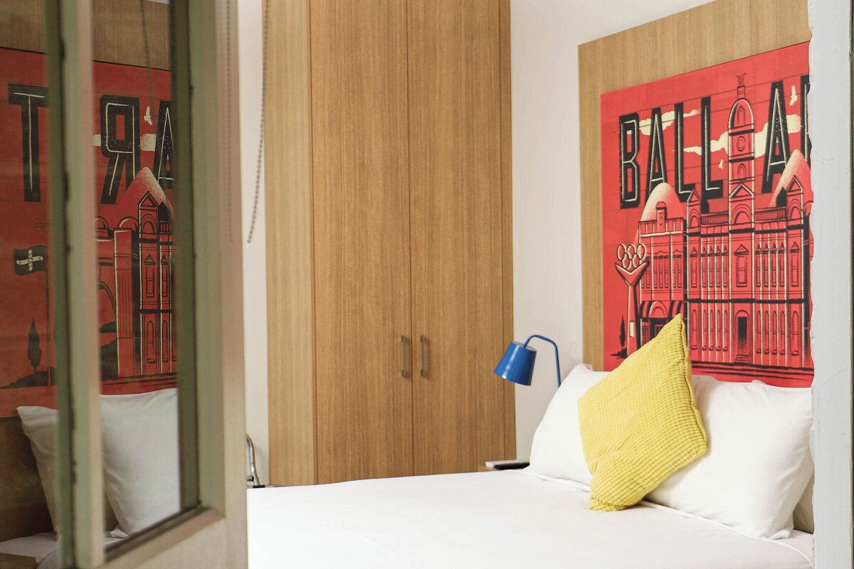 Une chambre à coucher avec un lit, une armoire en bois, un mur orné d’une fresque rouge et noire, et une lampe de chevet bleue.
